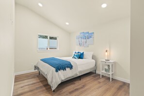 Un dormitorio con cama, ventana, mesita de noche con lámpara y pared decorada con tres cuadros.