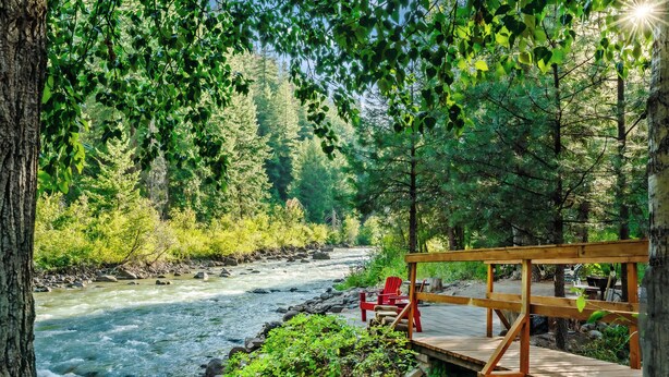 A wooden deck with red chairs overlooking a river, surrounded by dense forest.