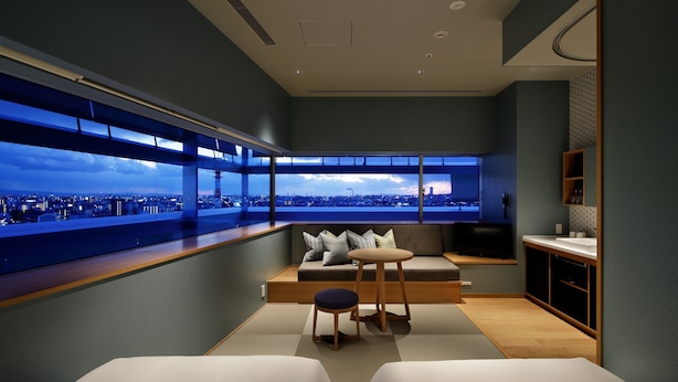 Ein modernes Hotelzimmer mit großem Fenster und Blick auf die Stadt, einer gemütlichen Sitzecke und einer kleinen Küchenzeile.