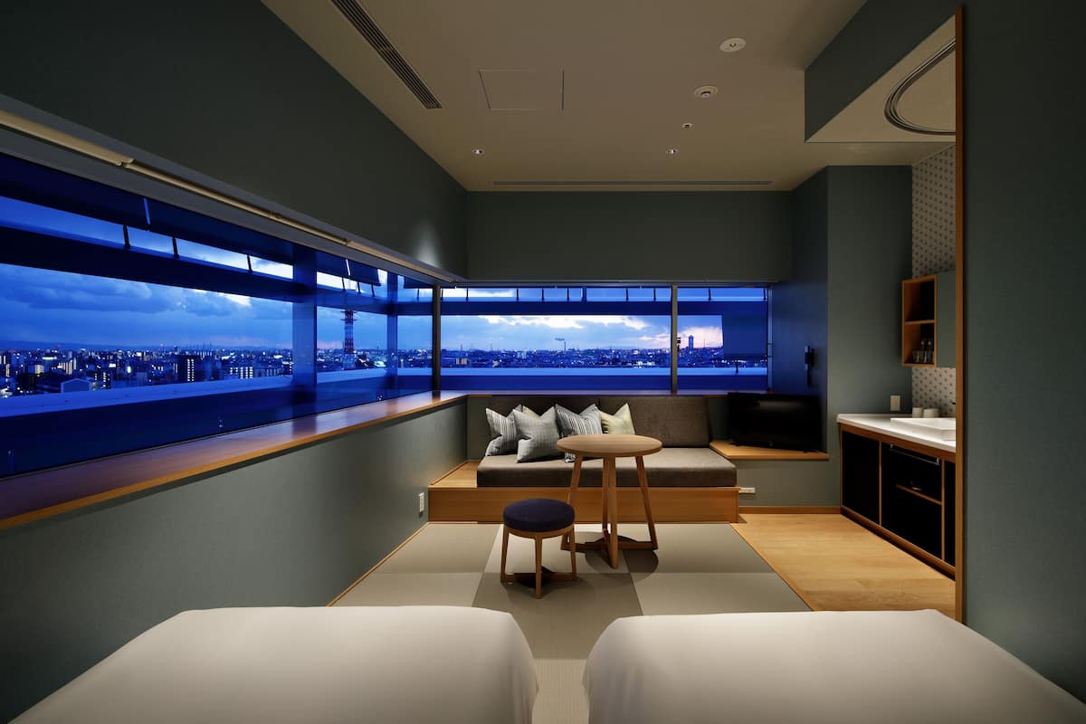 Ein modernes Hotelzimmer mit großem Fenster und Blick auf die Stadt, einer gemütlichen Sitzecke und einer kleinen Küchenzeile.