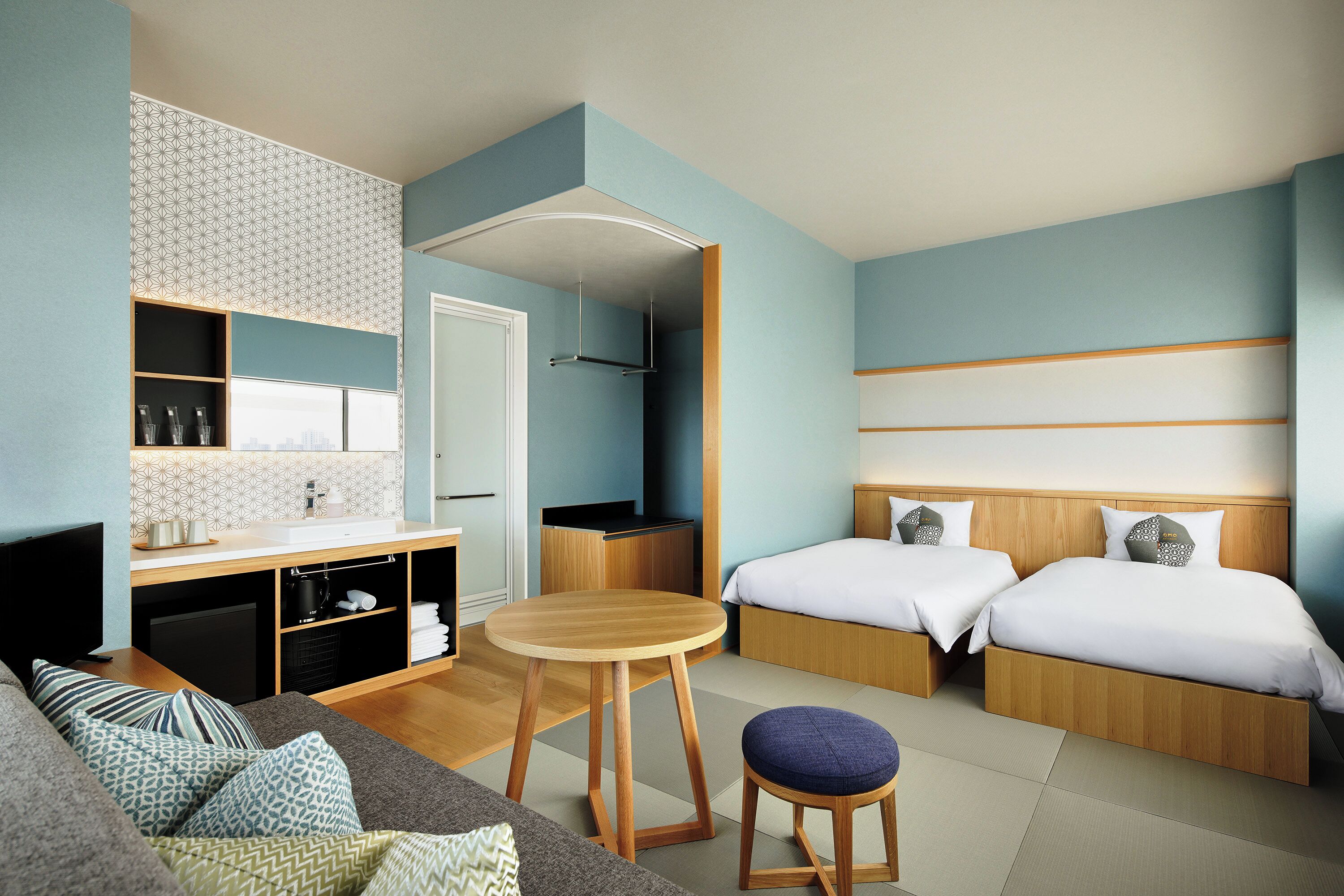 Una habitación de hotel moderna con kitchenette, un sofá, una mesa redonda de madera y dos camas.