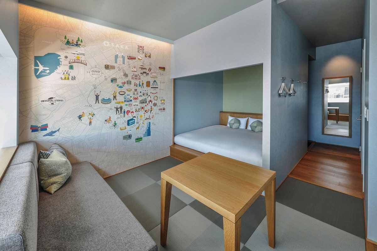 Ein modernes Hotelzimmer mit einer großen Wandkarte, einer Couch, einem Bett mit Kopfteil, einem Holztisch und einem kleinen Badezimmer mit Spiegel und Handtuchhalter.