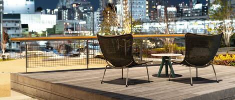 A wooden deck with two wicker chairs and a small table, overlooking a cityscape at night.