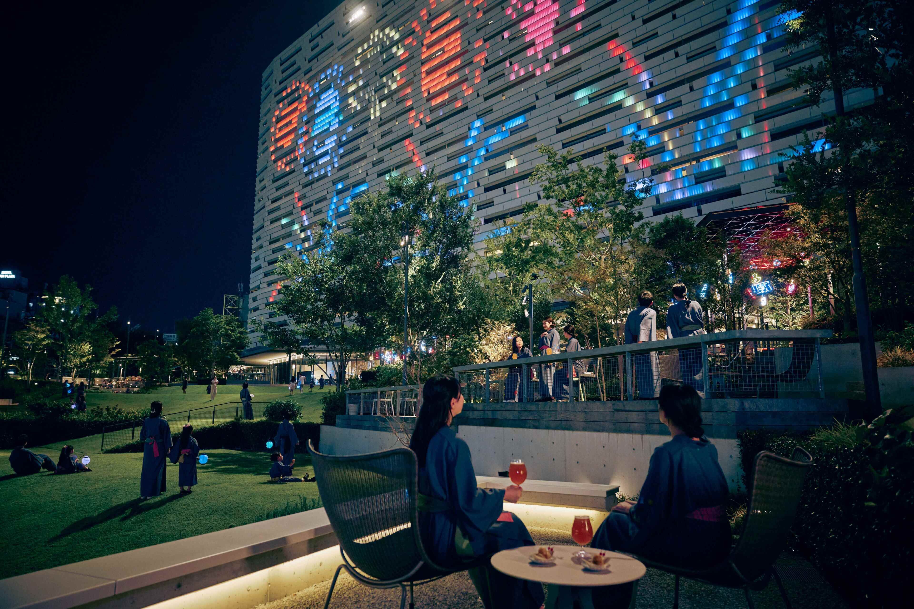 An outdoor seating area with tables and chairs, a modern building with illuminated windows in the background, and a well-maintained grassy area.
