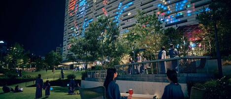 An outdoor seating area with tables and chairs, a modern building with illuminated windows in the background, and a well-maintained grassy area.