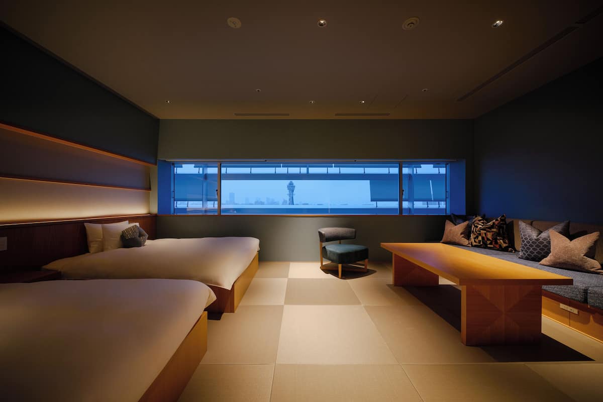 Ein modernes Hotelzimmer mit zwei Betten, einer Sitzecke, einem kleinen Tisch und Blick auf die Stadt durch große Fenster.