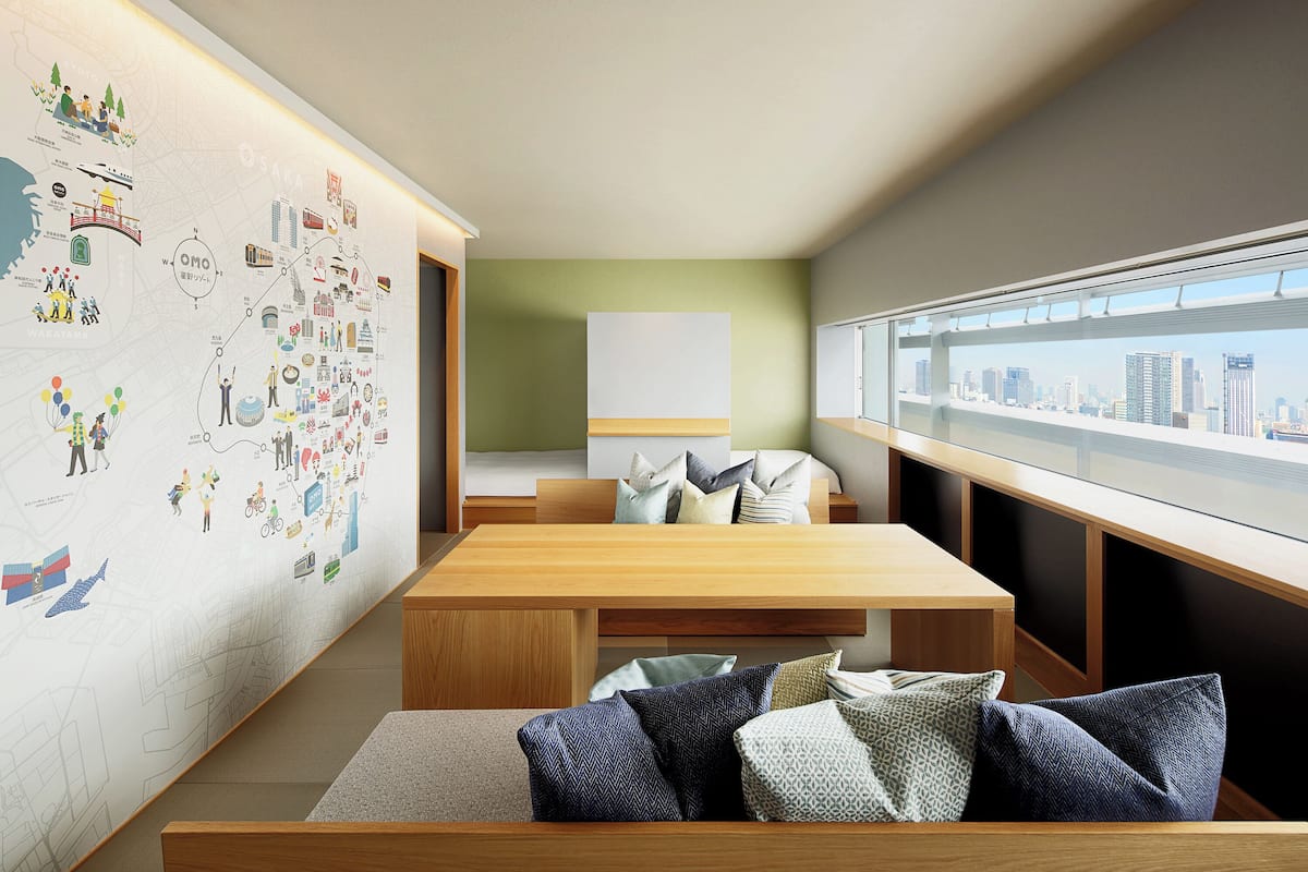 Ein modernes Hotelzimmer mit großem Fenster, einem Holzbüro, einer Couch mit gemusterten Kissen und einer Wand, die mit verschiedenen Illustrationen verziert ist.