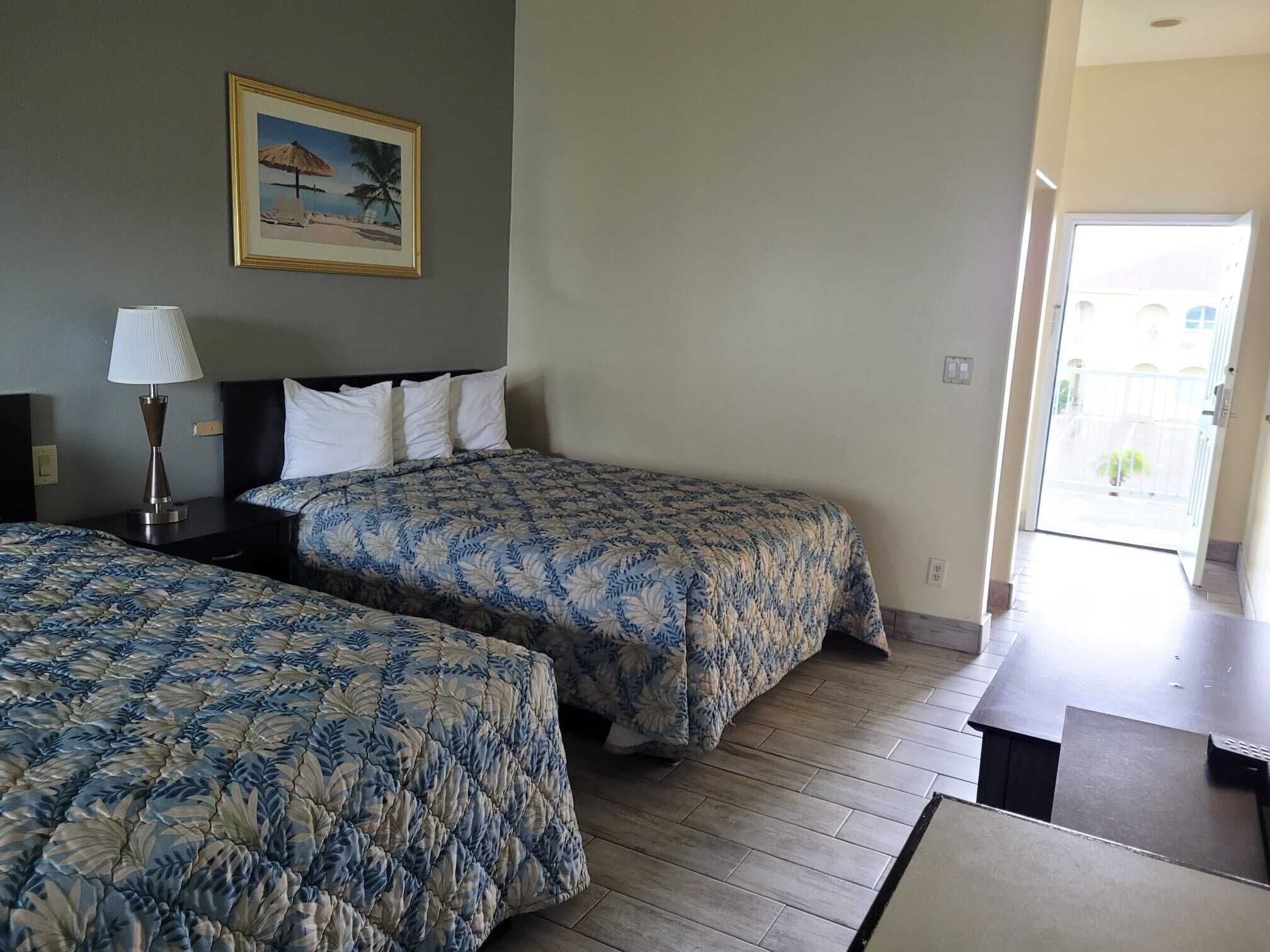 Habitación de hotel con dos camas, una mesita de noche con lámpara, un cuadro en la pared y vista al exterior a través de una puerta abierta.