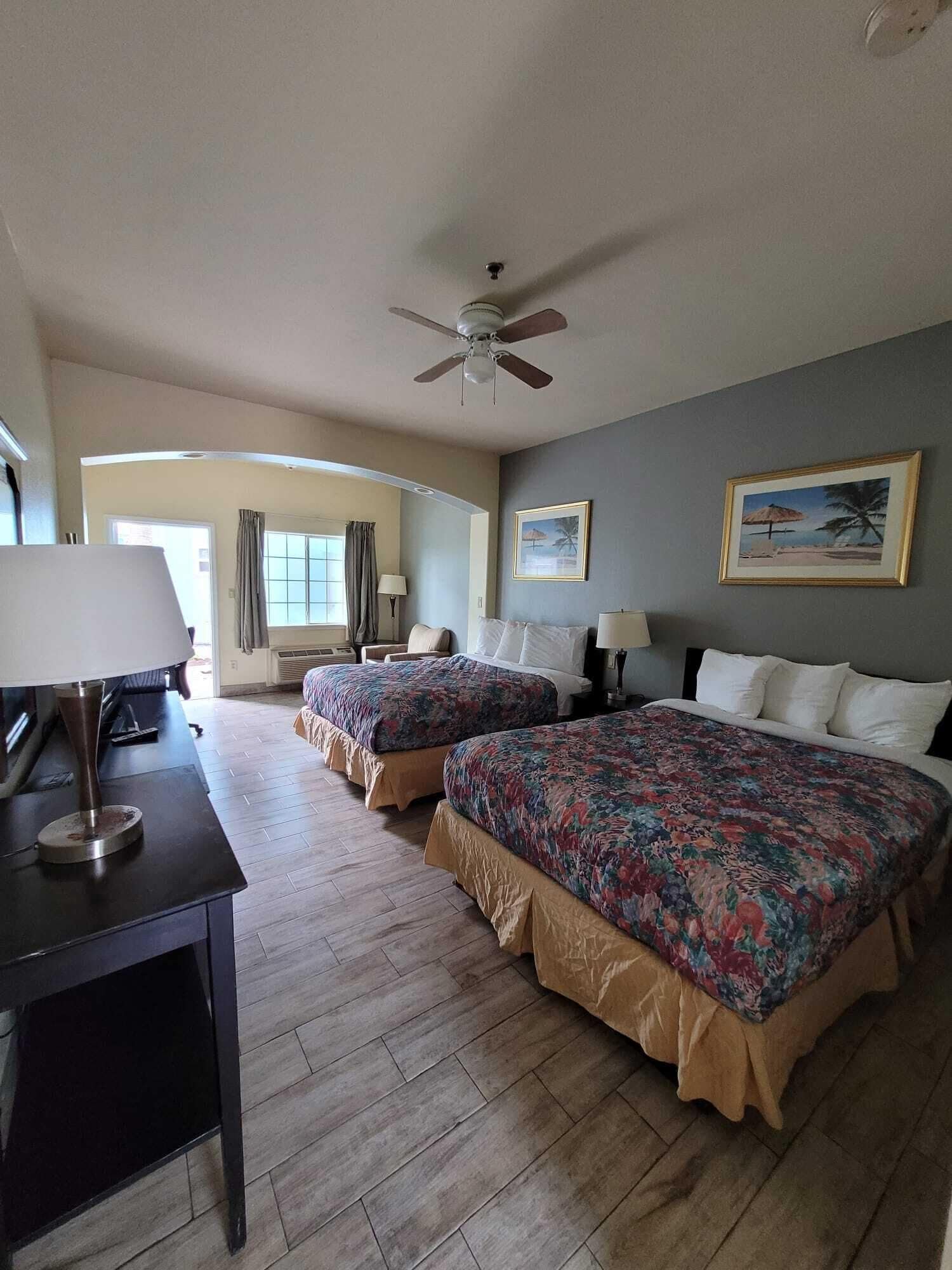 A hotel room with two beds, a desk with a lamp, a ceiling fan, and two framed pictures on the wall.