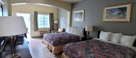 A hotel room with two beds, a desk with a lamp, a ceiling fan, and two framed pictures on the wall.