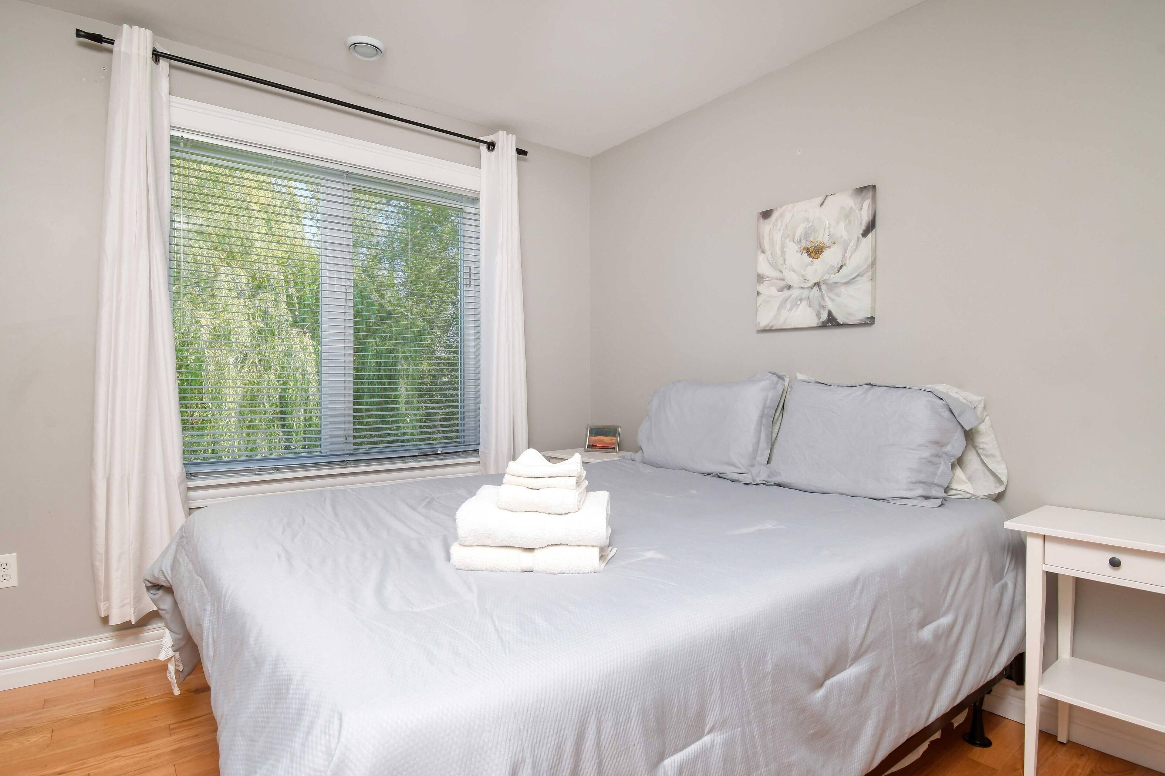 A bedroom with a bed, a nightstand, a window with blinds, and a wall-mounted artwork.