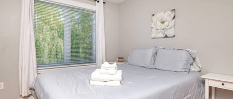 A bedroom with a bed, a nightstand, a window with blinds, and a wall-mounted artwork.