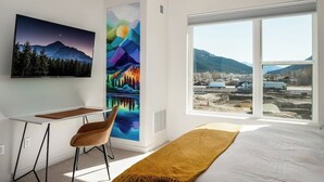 Un dormitorio con cama, escritorio, silla, un ventanal con vistas a las montañas y dos cuadros en la pared.