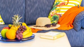 Ein blaues Sofa mit einem gestreiften Kissen und einem farbenfrohen Blumenkissen. Auf dem Sofa liegen ein Strohhut und eine Sonnenbrille. Eine Schale mit Obst, darunter Ananas, Trauben und Bananen, steht ebenfalls auf dem Sofa. Auf dem Tisch vor dem Sofa liegt ein Buch.
