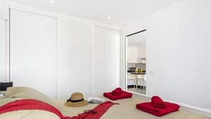 A modern bedroom with a large bed, a wardrobe, and a view of a kitchen through a sliding door.