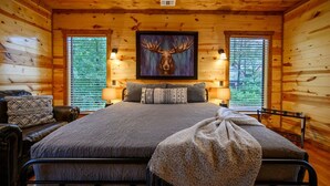Dormitorio de cabaña de madera con cama, dos lámparas, cabeza de alce enmarcada y ventana con persianas.