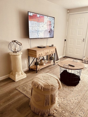 Ett vardagsrum med en tv som är monterad på väggen, ett soffbord i trä, en rund pall, ett litet sidobord med dekorativa föremål och en mönstrad matta.