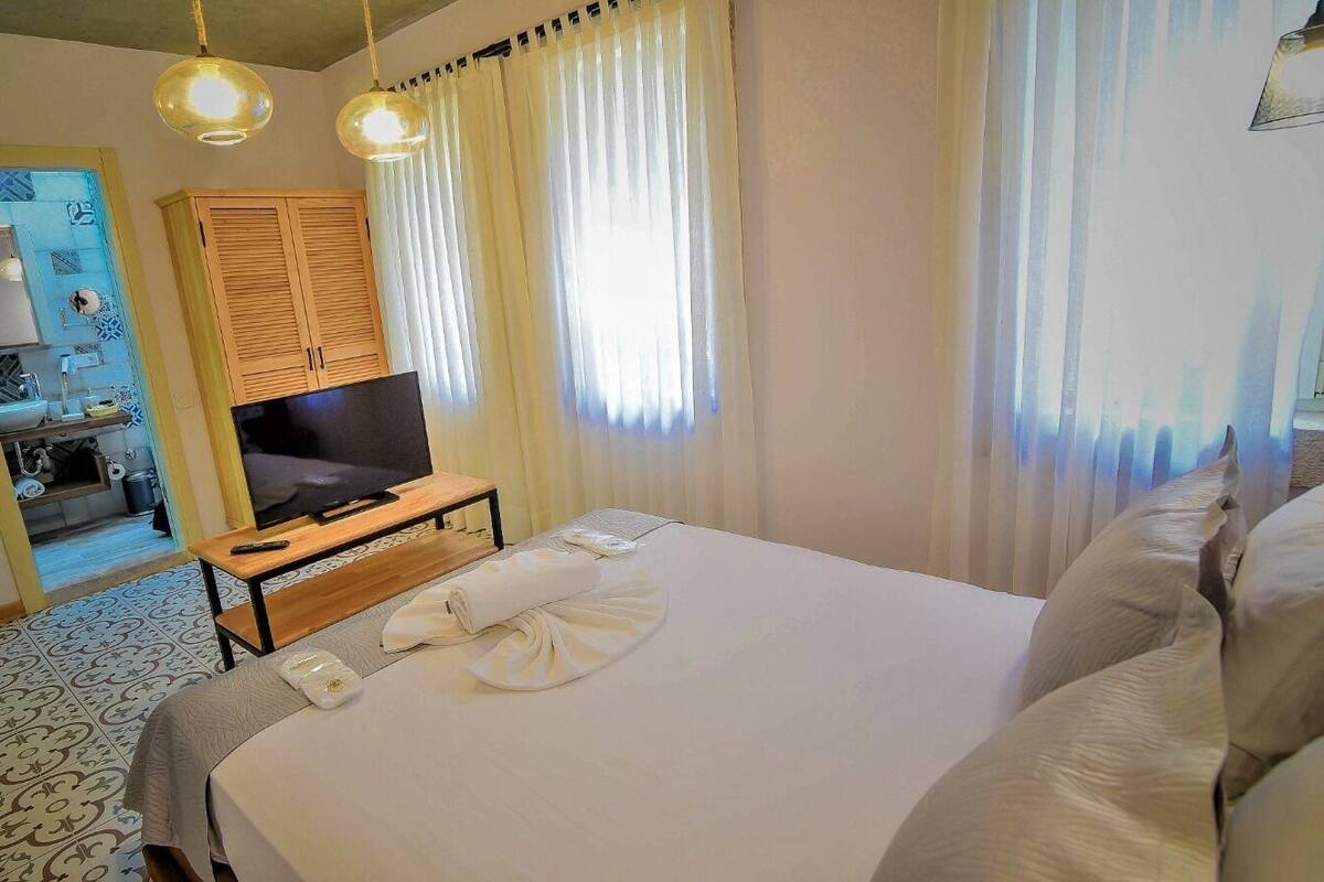 A hotel room with a large bed, a television on a wooden stand, a bathroom with a mirror and sink, and a view of the outside through sheer curtains.