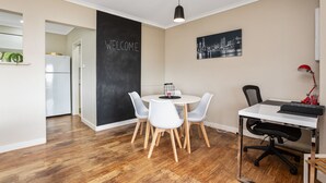 A modern kitchen with a white refrigerator, a blackboard with "WELCOME" written on it, a dining table with white chairs, and a cityscape painting on the wall. - Beach Haven on The Hill (The Hill)