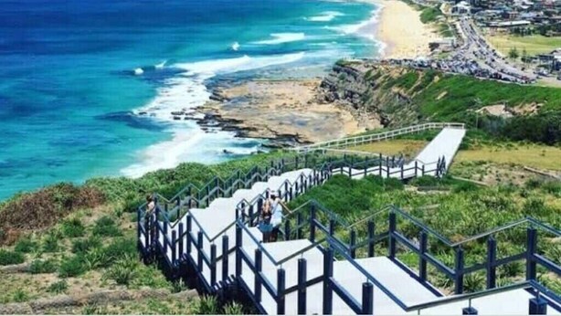 A coastal town with a staircase leading down to the beach, a clear view of the ocean, and a road running parallel to the coastline. - Beach Haven on The Hill (The Hill)