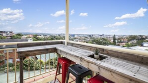 A balcony with a wooden table, two red barstools, and a potted plant. - Beach Haven on The Hill (The Hill)