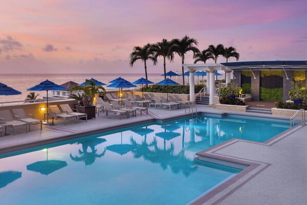 A pool area with lounge chairs and umbrellas, a clear view of the ocean, and a sunset sky.