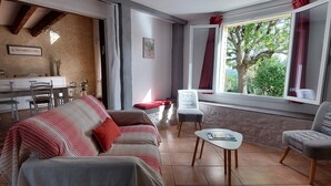 A cozy living room with a sofa, a small table, and a chair. There is a dining area with a table and chairs. A window with curtains is visible, and a plant is on the windowsill. - Le Bellevue, quiet gite with panoramic view 10 minutes from Sarlat (Vezac)