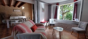 A cozy living room with a sofa, a small table, and a chair. There is a dining area with a table and chairs. A window with curtains is visible, and a plant is on the windowsill. - Le Bellevue, quiet gite with panoramic view 10 minutes from Sarlat (Vezac)