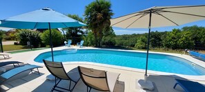 A swimming pool with lounge chairs and umbrellas, surrounded by greenery and a fence.