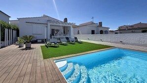 Uma área à beira da piscina com um deck de madeira, uma piscina e um edifício branco com um terraço.