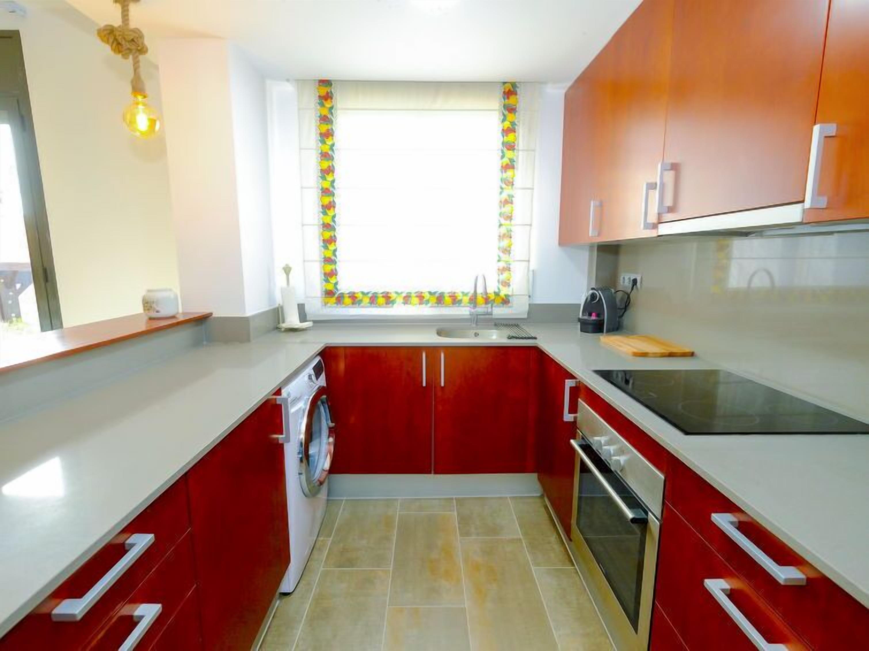 A compact kitchen with red cabinets, a built-in oven, and a window with a colorful curtain.