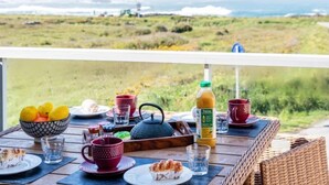 Een ontbijttafel met diverse spullen, zoals een fruitschaal, kopjes, een theepot en een fles sap, op een balkon met uitzicht op een grasland en de zee.