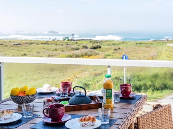 A breakfast table with various items including a bowl of fruit, cups, a teapot, and a bottle of juice, set on a balcony overlooking a grassy landscape and the sea.