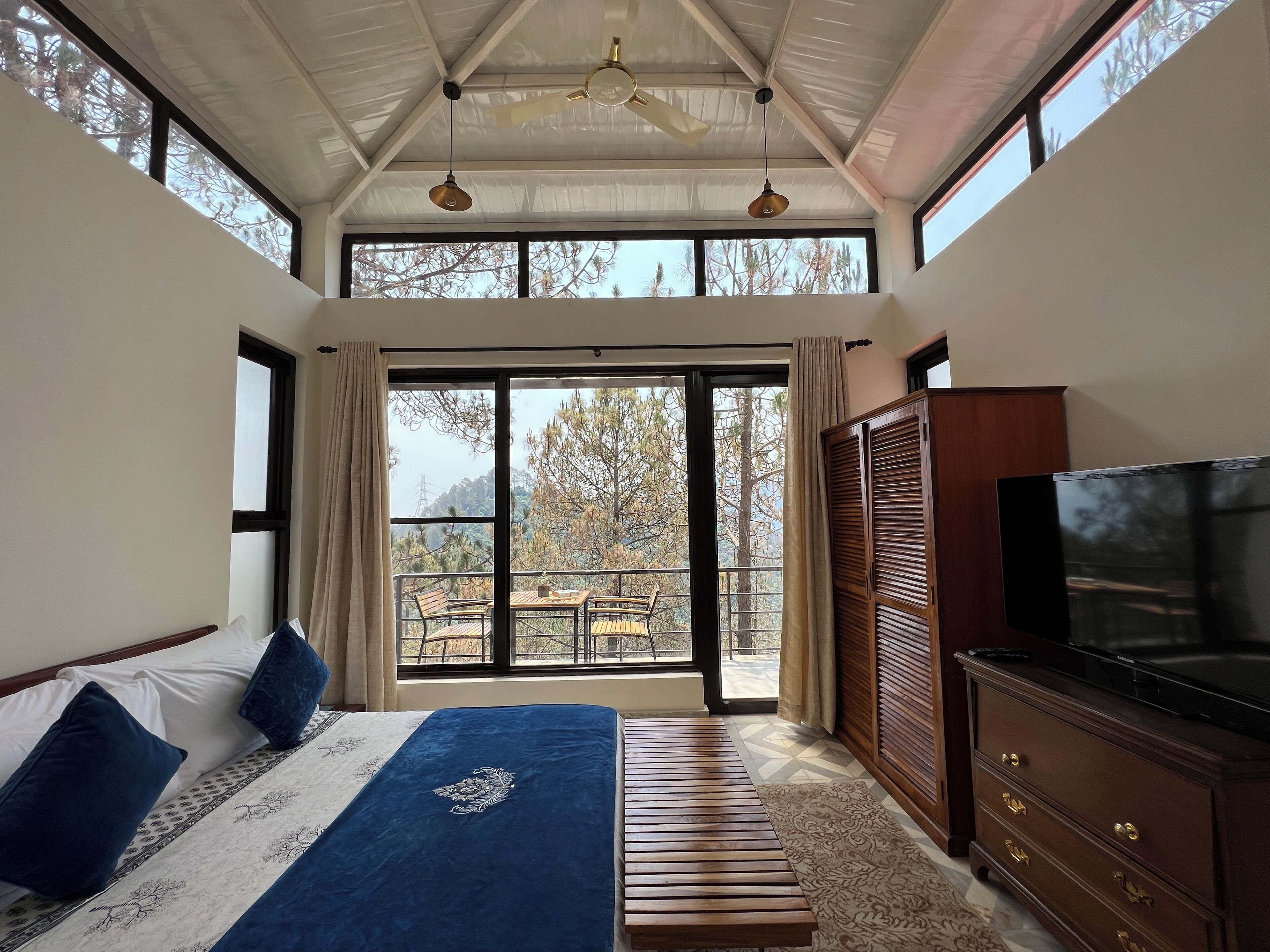 A bedroom with a bed, a TV, a dresser, and a balcony with a view of trees.