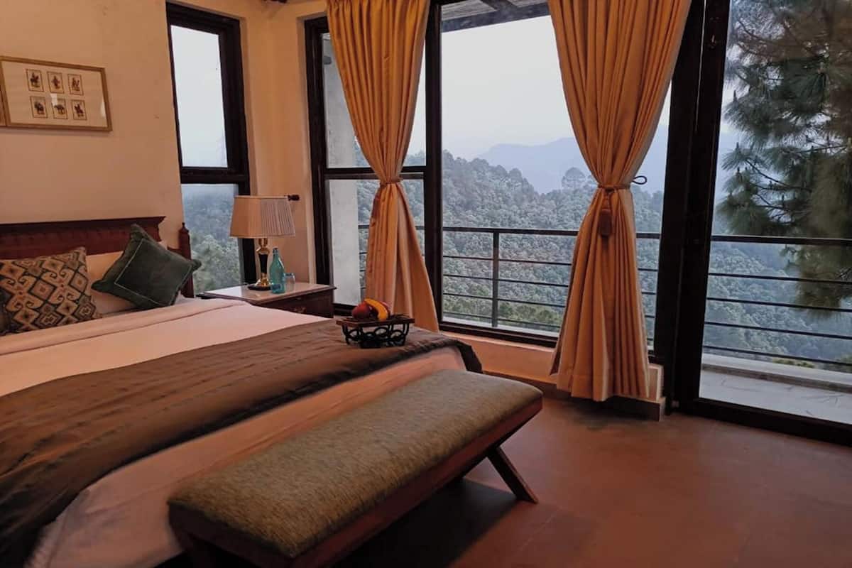 A bedroom with a large bed, a bench, a nightstand with a lamp, and a view of a mountainous landscape through large windows.