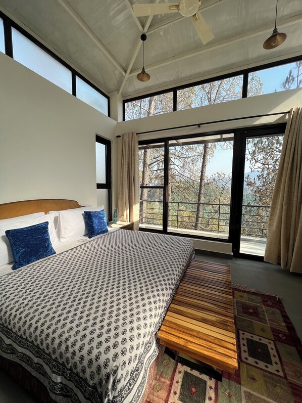 Un dormitorio con una cama grande, un banco de madera y vistas a árboles a través de ventanas amplias.