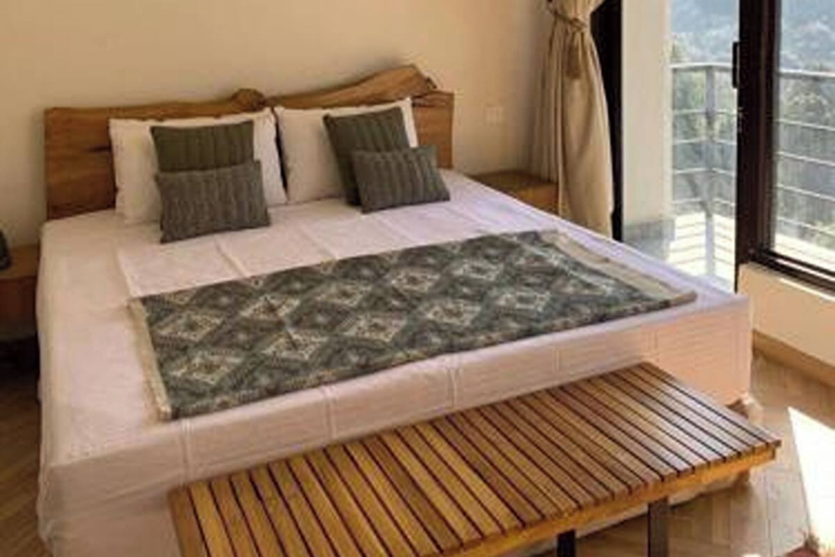 A bedroom with a bed, wooden headboard, two bedside lamps, a window with curtains, and a view of the outdoors.