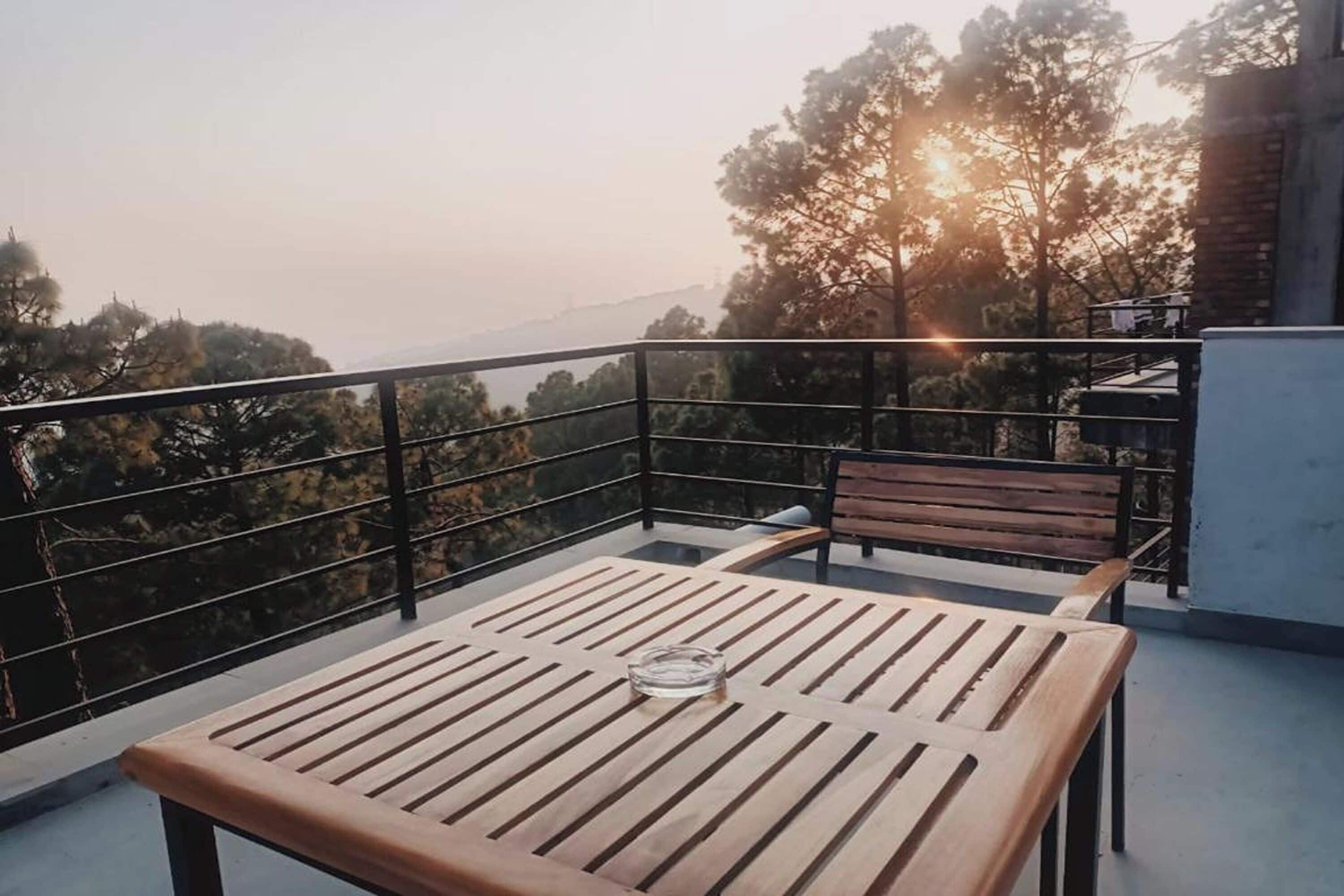 Un balcón con mesa y banco de madera, con vista a una zona boscosa llena de árboles y una lejana vista de colinas.