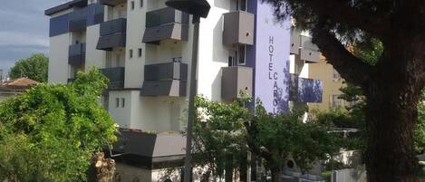 A multi-story hotel with balconies, a sign reading "HOTEL CARLO," and surrounding greenery.