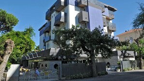 Un bâtiment moderne à plusieurs étages, doté de balcons, d’un espace extérieur aménagé et couvert, et d’un arbre au premier plan.