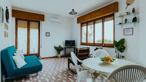 A cozy living room with a sofa, a dining table with chairs, a TV, and a large window with wooden shutters. - Heart of Ognina - Veliero - Apartment with sea view (Ognina)