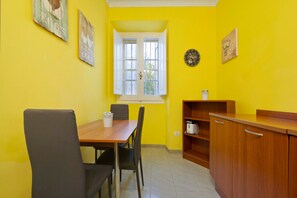 A room with a dining table, chairs, a window, a clock, and a kitchen area with a cabinet and a toaster.