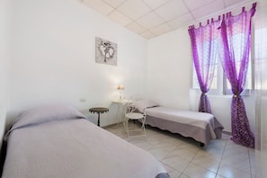A room with two beds, a small table, a chair, a lamp, and a window with purple curtains.
