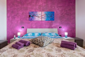 Ein Hotelzimmer mit einer rosafarbenen, strukturierten Wand, einem Bett mit floralem Bettbezug, zwei Nachttischen mit Lampen und einem Baumgemälde.