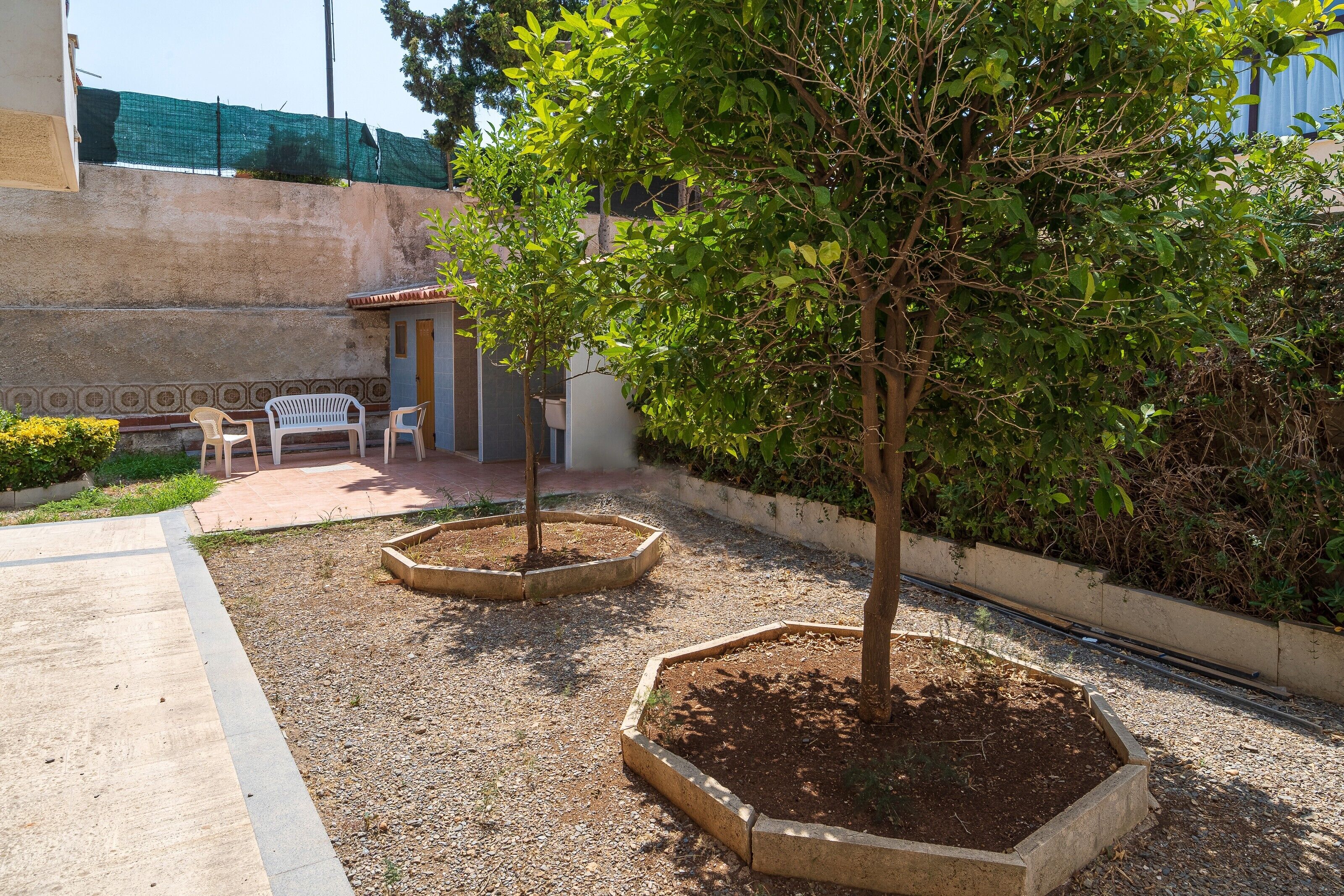 Ein Innenhof mit zwei Bäumen in erhöhten Blumenbeeten, einer kleinen Sitzgruppe mit Stühlen und einem Betonweg.