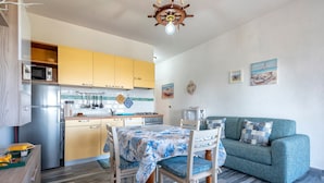 Et kompakt køkken med moderne hårde hvidevarer, et spisebord med stole, og en turkis sofa i et lyst opholdsrum.
