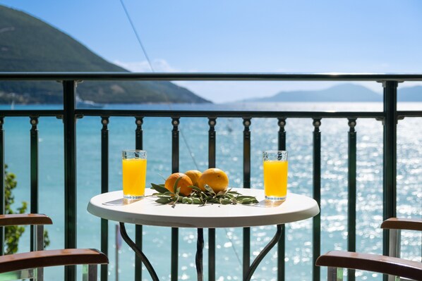 A table with two glasses of orange juice and three oranges on a balcony overlooking a body of water.