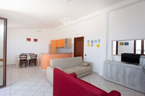 Ein Wohnzimmer mit Sofa, Fernseher, Esstisch mit Stühlen und einer Küchenzeile im Hintergrund.