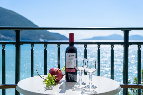 Une table avec une bouteille de vin, deux verres et une grenade, sur un balcon offrant une vue sur la mer et les montagnes.