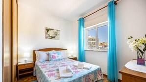 A bedroom with a bed, nightstands, a window with curtains, a painting, and a desk with a vase of flowers. - Apartment Villas Brites nº4 with Shared Pool, Shared Garden, Balcony & Wi-Fi (Burgau)